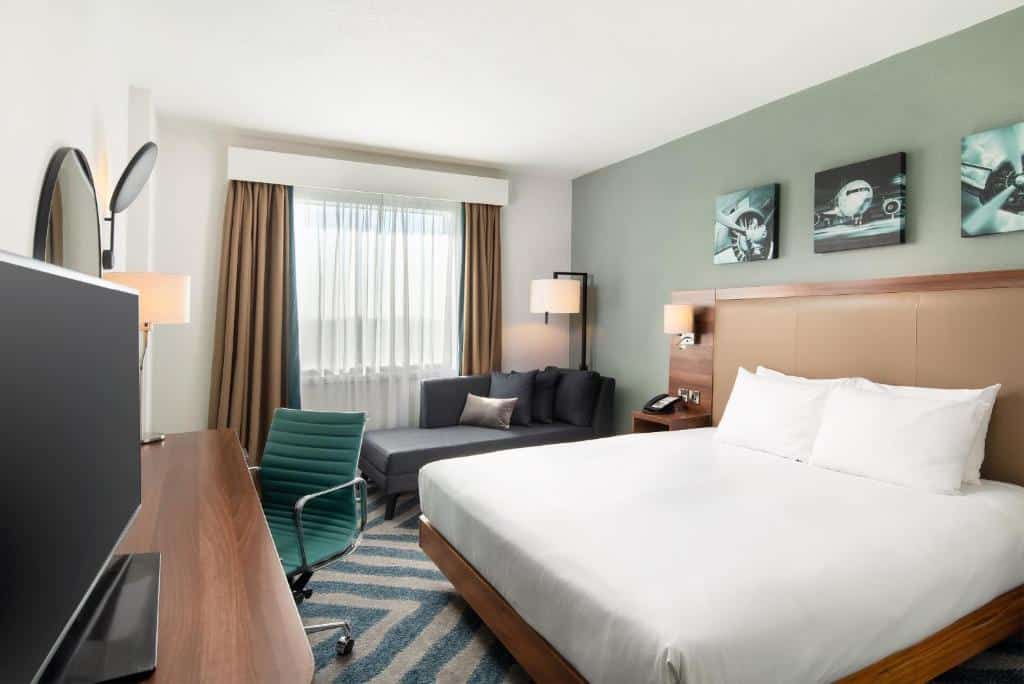 Quarto do Hilton Garden Inn London Heathrow Airport com uma cama de casal, um carpete azul e branco, uma janela com cortinas, uma mesa com uma televisão e uma cadeira giratória