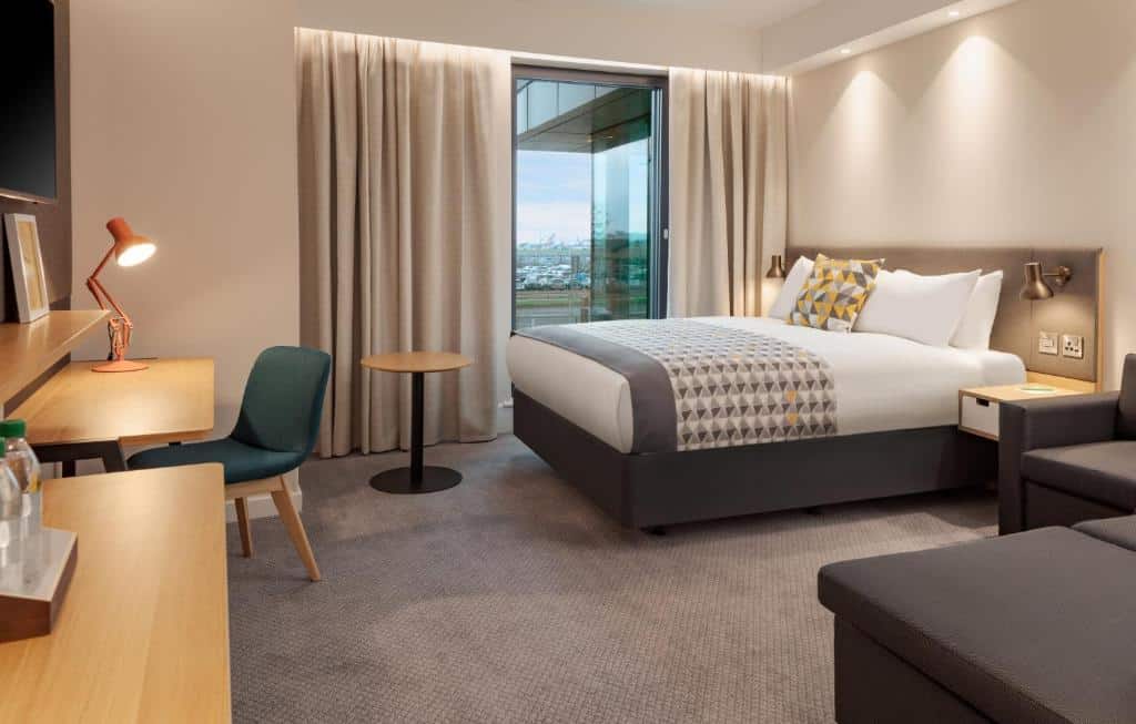 Quarto do Holiday Inn London Heathrow - Bath Road, an IHG Hotel com uma cama de casal, uma mesa com uma cadeira e uma luminária, tem um sofá também em um dos cantos, para representar hotéis perto do Aeroporto de Londres Heathrow