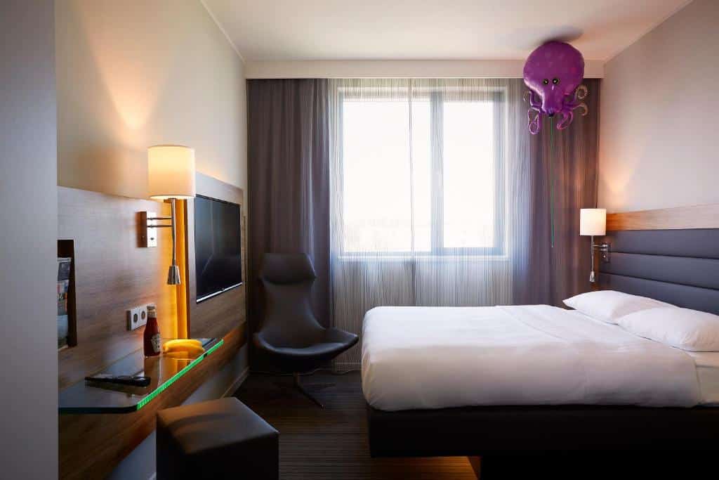Quarto do Moxy London Heathrow Airport com uma cama de casal, uma janela com cortinas, chão de madeira, uma televisão de frente para a cama, e uma poltrona