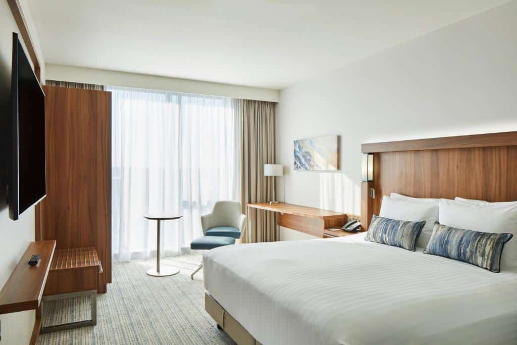 Quarto do Courtyard by Marriott London Heathrow Airport com uma janela com cortinas, uma cama de casal, uma televisão, um armário, uma mesinha com um abajur e uma poltrona, para representar hotéis perto do Aeroporto de Londres Heathrow