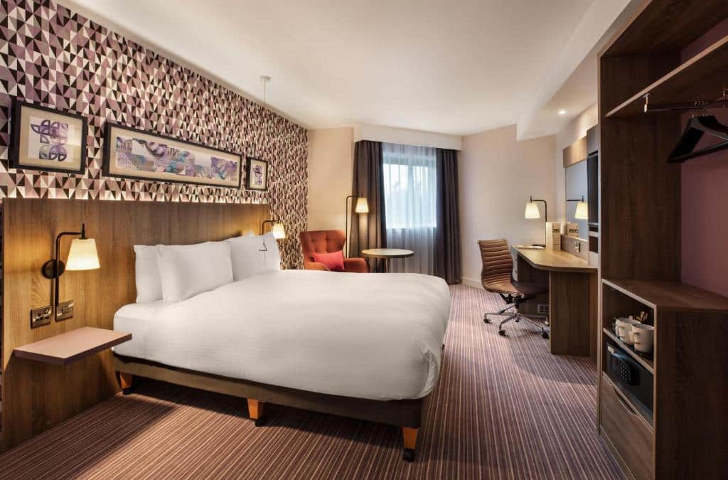 Quarto do Leonardo London Heathrow Airport com uma janela com cortinas, uma cama de casal, um armário conceito aberto, um carpete, uma mesinha de escritório com uma cadeira giratória, uma poltrona e um abajur de chão, para representar hotéis perto do Aeroporto de Londres Heathrow