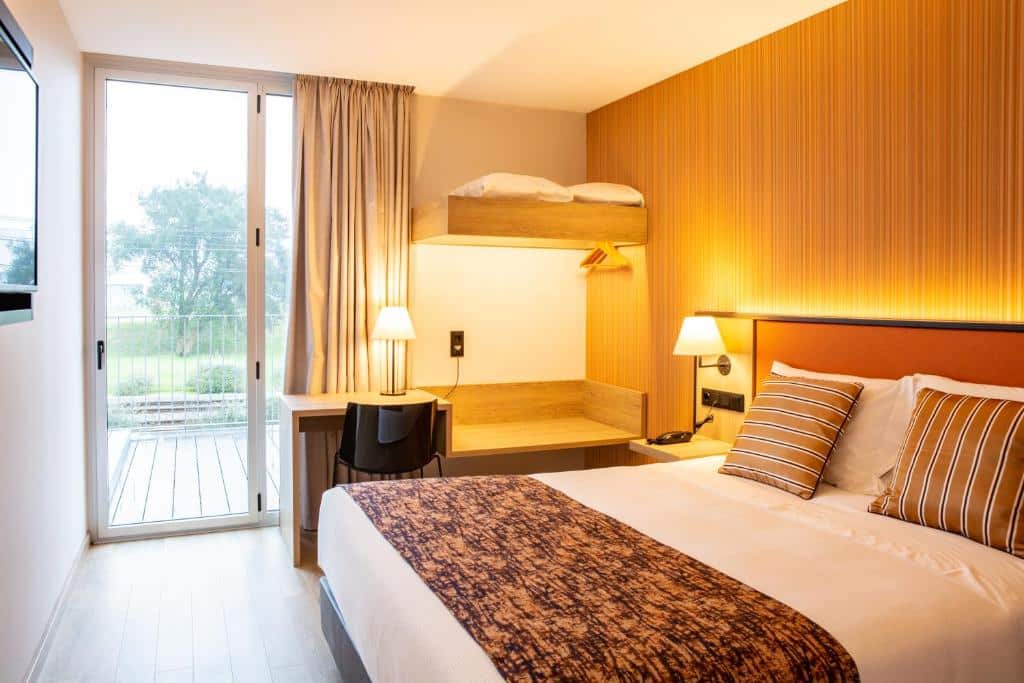 Quarto do Hotel 3K Porto Aeroporto com cama de casal do lado direito, uma TV em frente a cama presa na parede, do lado esquerdo da cama uma mesa de trabalho com cadeira. Representa hotéis perto do aeroporto do Porto.
