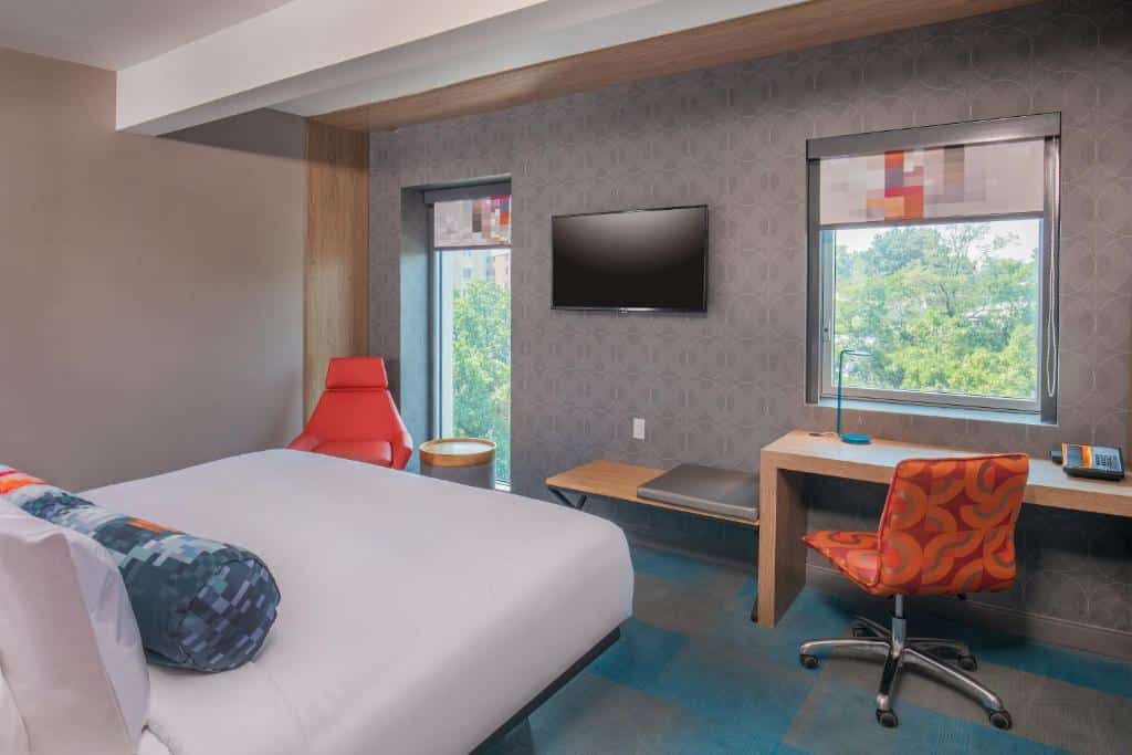 quarto do Aloft New York LaGuardia Aeroporto com uma cama de casal à esquerda de frente para a televisão de tela plana presa a parede, entre duas janelas com vista para árvores verdes