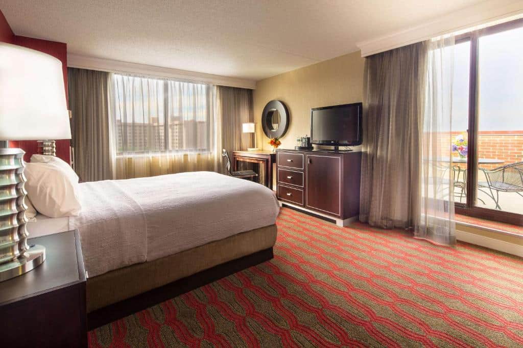 suíte do LaGuardia Plaza Hotel com uma cama de casal do lado esquerdo, disposto em frente a televisão de tela plana sobre uma cômoda de madeira, ao lado da porta de vidro que dá acesso ao terraço privativo, onde há uma mesa e duas cadeiras de ferro.