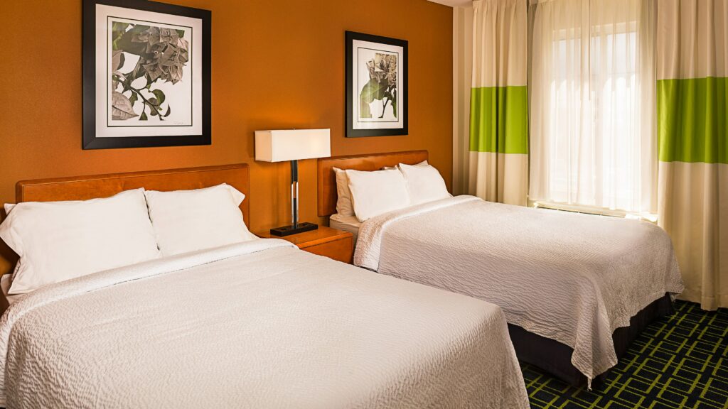 suíte do Fairfield Inn Flushing, um dos hotéis perto do aeroporto laguardia em nova york, com duas camas de casal dispostas lado a lado. Em cima de cada cama há um quadro com imagens de flores.
