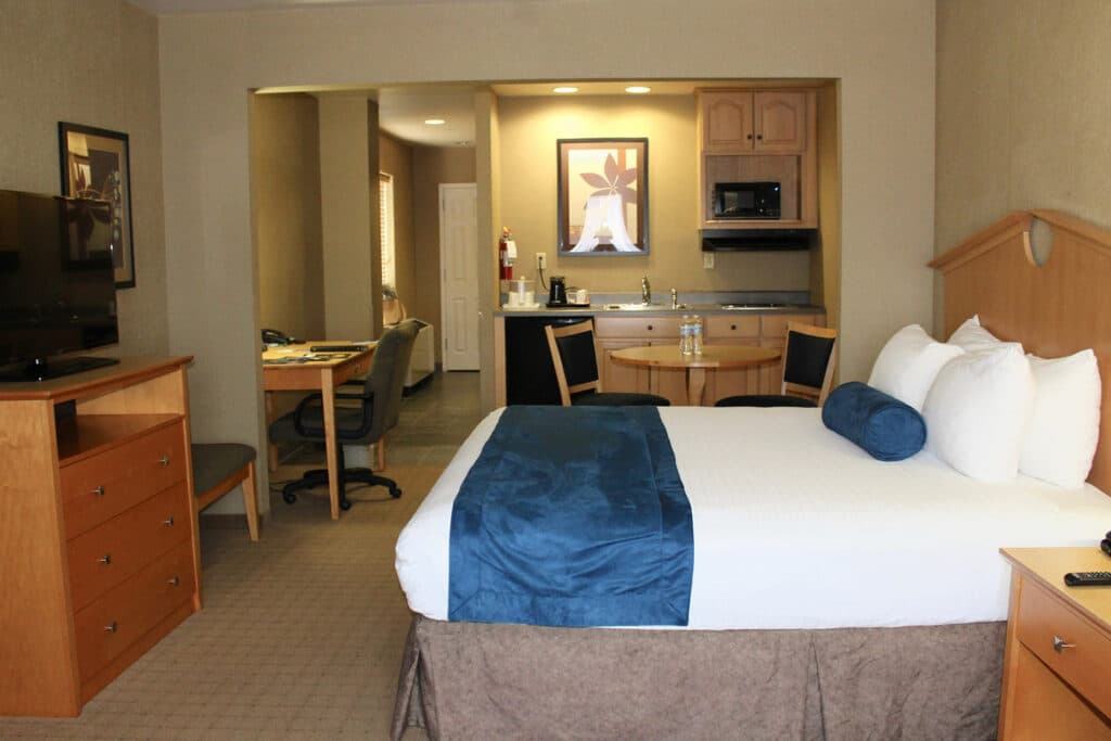 o quarto do Marco LaGuardia Hotel & Suites é mais um dos hotéis perto do aeroporto laguardia em nova york com uma cama de casal à direita, em frente a uma cômoda com televisão em cima. Mais ao fundo quarto é possível ver uma pequena cozinha com uma mesa redonda, duas cadeiras e uma mesa retangular à esquerda do quarto, com uma cadeira estofada para trabalho.
