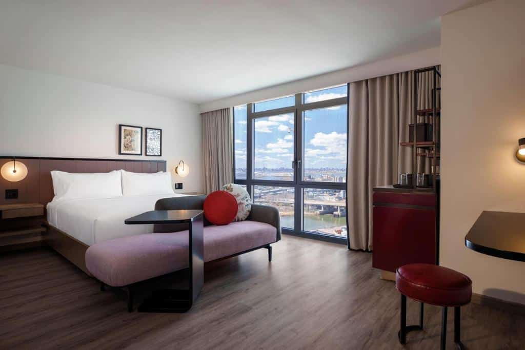 suíte elegante do Renaissance New York Flushing Hotel at Tangram, um dos hotéis perto do aeroporto laguardia em nova york, com uma cama de casal e um sofá ao pé da cama no lado esquerdo, uma parede coberta de janelas com vista para a cidade ao fundo e uma prateleira de madeira à direita com um frigobar vermelho