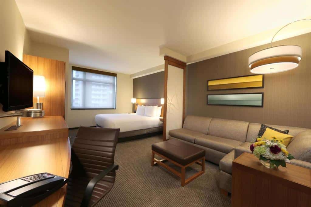 quarto do Hyatt Place Flushing/LGA Airport com uma cama de casal ao fundo, separada por uma pequena divisória de ambientes da sala de estar com um sofá de canto cinza, em frente a mesa comprida de madeira para trabalho, com uma televisão sobre ela.