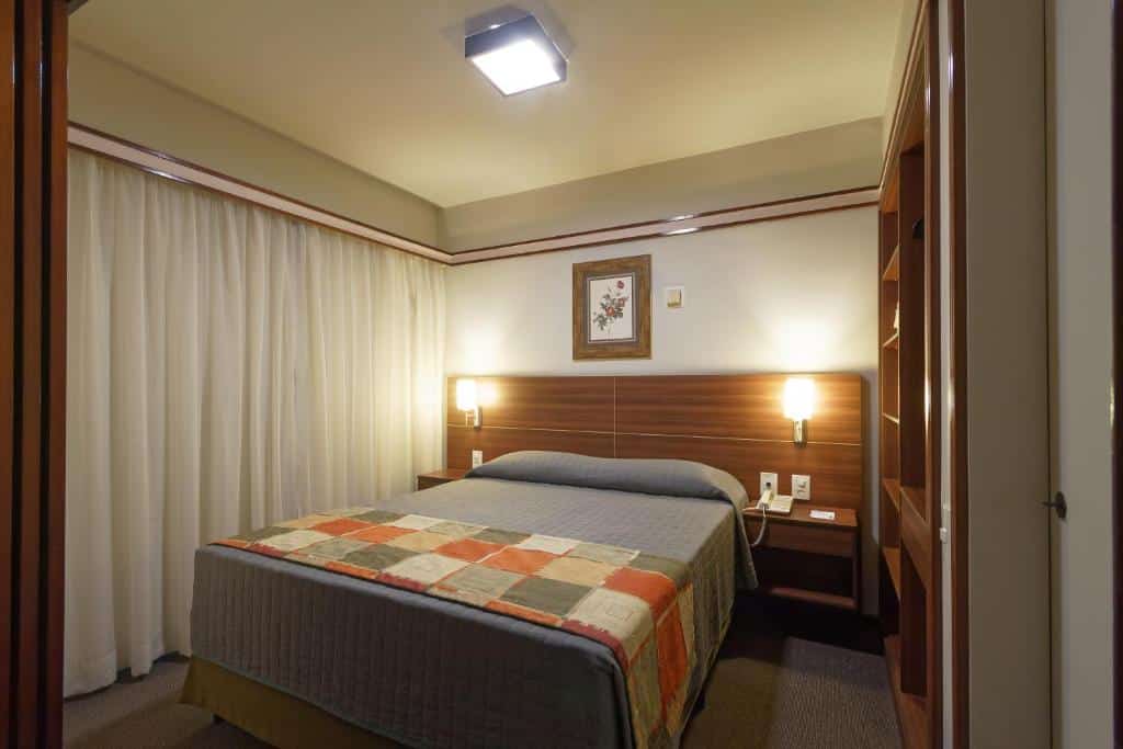 Quarto no Bristol The Time Berrini com uma cama de casal, uma janela do lado esquerdo, do lado direito há um armário de conceito aberto, a cabeceira tem dois abajures presos e o chão é um carpete cinza
