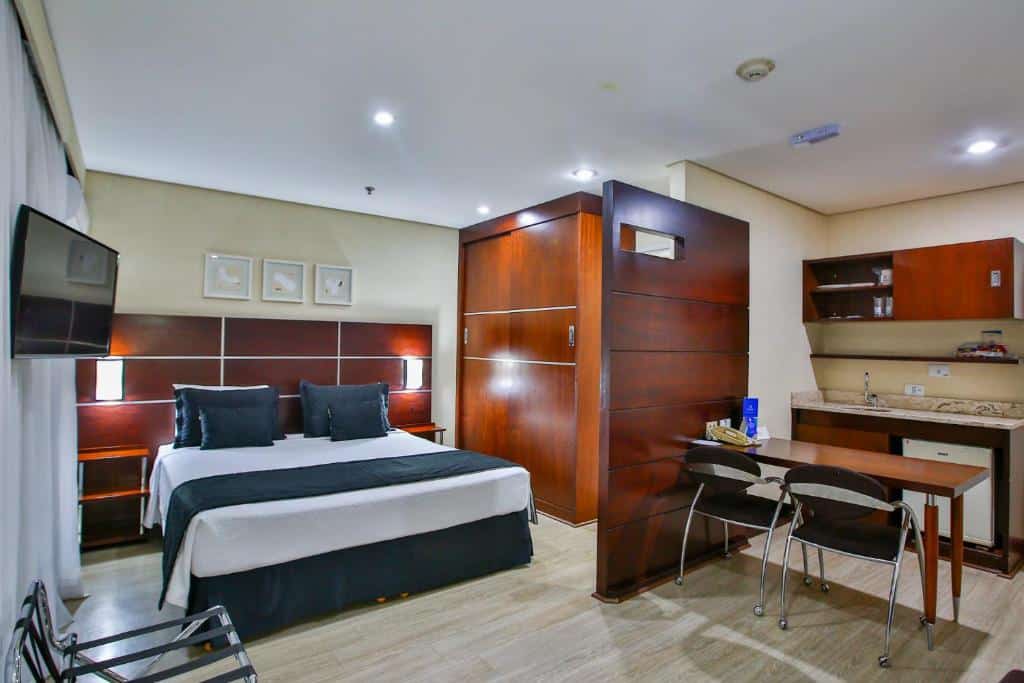 Quarto do Transamerica Executive Chácara Santo Antônio com uma cozinha compacta, uma mesa com dois lugares, uma cama de casal e uma televisão, o chão é de madeira e há uma janela do lado esquerdo com cortinas, para representar os hotéis perto do Autódromo de Interlagos