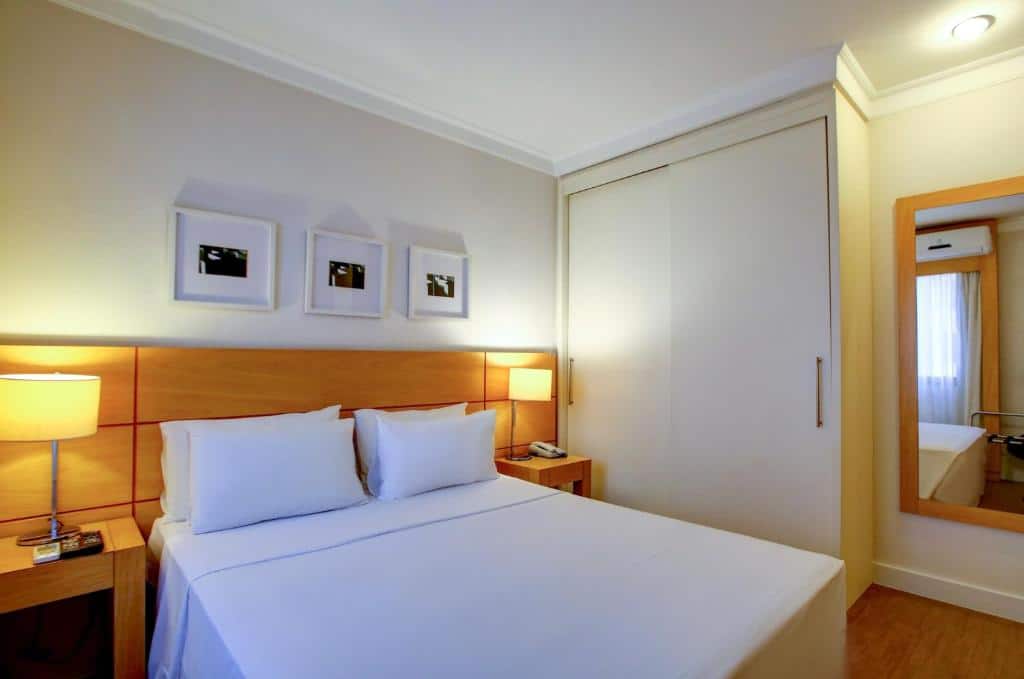Quarto do eSuítes Congonhas by Atlantica com um pequeno armário, um espelho, uma cama de casal com duas mesinhas de cabeceira com luminárias presas nelas, há três quadros pequenos sob a cama e o chão é de madeira clara