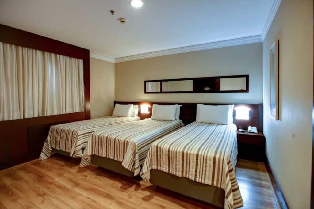 Quarto no Transamerica Executive Congonhas com três camas de solteiro, do lado esquerdo tem uma janela com cortinas, entre as camas há abajures presos na cabeceira e o chão é de madeira