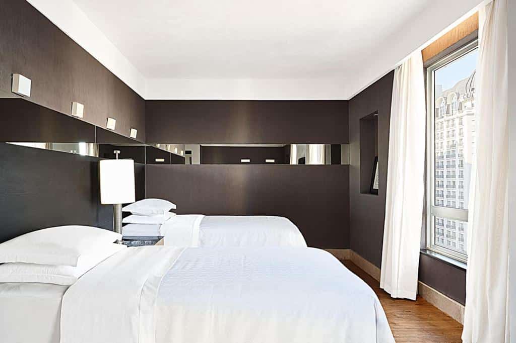 Quarto no Sheraton São Paulo WTC Hotel com duas camas de solteiro viradas para uma ampla janela com cortinas, o chão é de madeira e há um abajur entre as duas camas, para representar hotéis perto do Autódromo de Interlagos