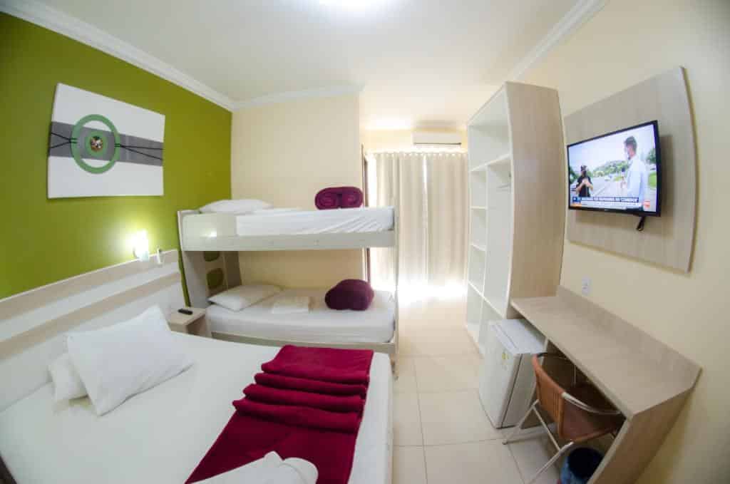 Quarto com uma cama de casal, um beliche, uma TV, uma mesa, uma cadeira, um armário, um frigobar e um ar-condicionado em um dos hotéis perto do Beto Carrero World
