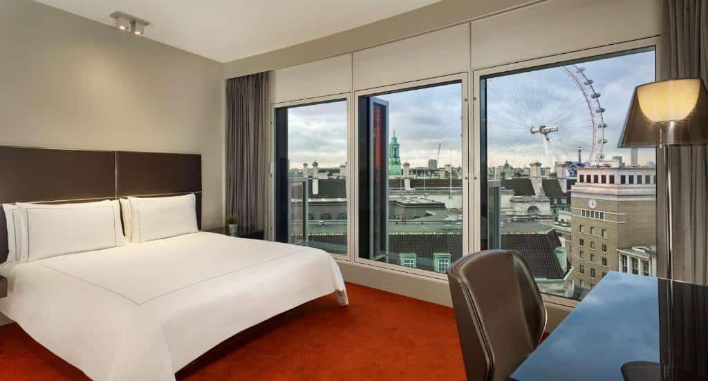 Quarto do Park Plaza Westminster Bridge London com três janelas grandes com vista para a cidade e a London Eye, uma cama de casal, carpete vermelho, uma mesa de escritório com uma cadeira, para representar hotéis perto do Big Ben em Londres