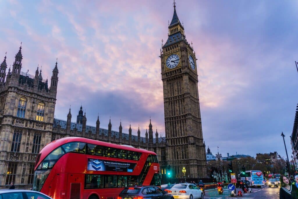 Prédio do Big Ben com a grande torre destacada em uma rua movimentada com ônibus vermelhos, carros e pessoas caminhando em um fim de tarde
