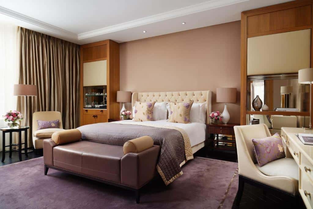Quarto do Corinthia London com uma cama de casal, uma tapete roxo, um pequeno sofá, uma penteadeira com uma cadeira e uma janela com cortinas