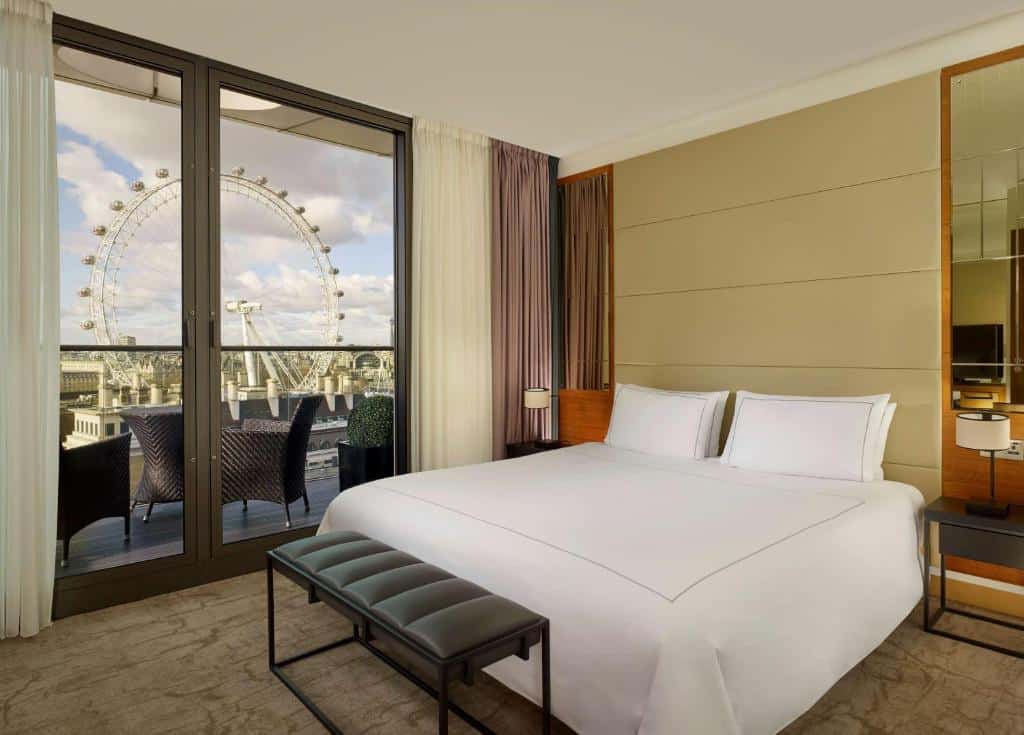 Quarto do Park Plaza County Hall London com uma varanda com cadeira e mesa com vista para a London Eye, com uma cama de casal, duas mesinhas de cabeceira e um pequeno banco, para representar hotéis perto do Big Ben em Londres