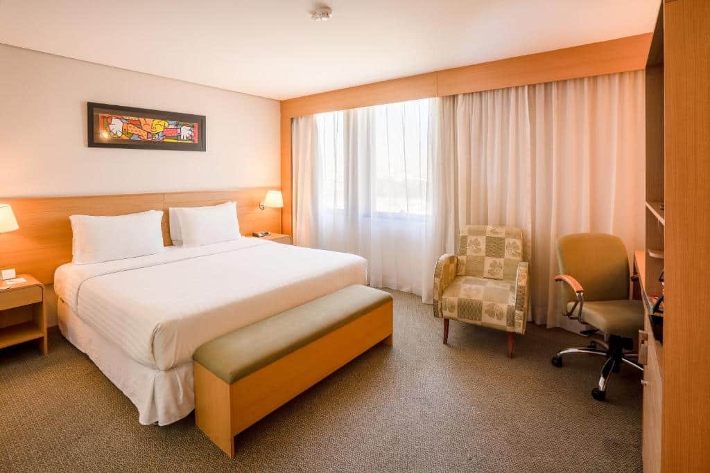 Quarto do Holiday Inn Parque Anhembi, an IHG Hotel. Uma mesa de trabalho e uma poltrona do lado direito. Do lado esquerdo uma cama de casal, com um banco de apoio e uma cômoda e um abajur de cada lado. No fundo a janela do quarto. Imagem para ilustrar post sobre hotéis perto do Expo Center Norte.