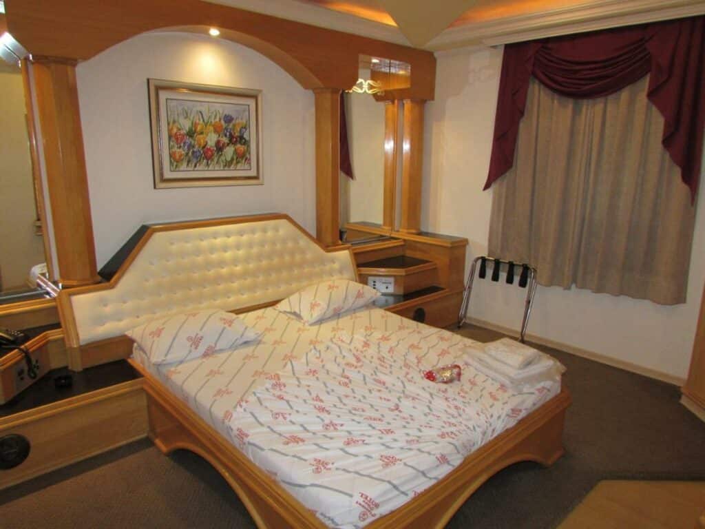 Quarto do Pleasant Place Hotel. Uma cama de casal no centro, de cada lado um espelho. No fundo uma janela com cortina.