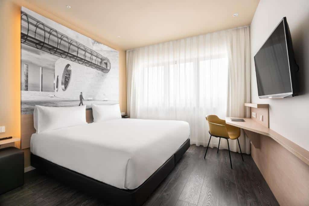 Quarto do Ikonik Lisboa com uma cama de casal, uma janela ampla com cortinas brancas, uma televisão de frente para a cama e uma pequena mesa de escirtório próxima da janela, junto de uma cadeira.