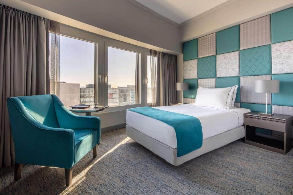 Quarto do Tivoli Oriente Lisboa Hotel com uma janela ampla com cortinas, uma cama de solteiro com uma cabeceira estofada em branco e azul claro, há duas mesinhas de cabeceira com abajures e uma poltrona estofada em azul perto da janela.