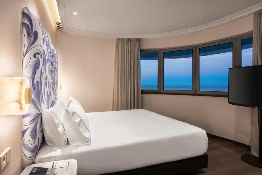 Quarto do Eurostars Universal Lisboa com uma cama de casal virada para janelas que dão vista para o Rio Tejo, há uma televisão e luminárias presas na parede ao lado da cama, para representar hotéis perto do Parque das Nações em Lisboa.