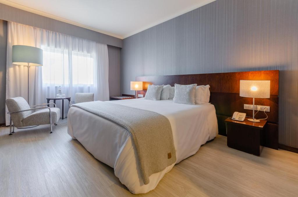 Quarto do Olissippo Oriente com uma cama de casal com travesseiros e almofadas, nas laterais da cama há mesinhas com abajures, do lado esquerdo do quarto há uma janela com cortinas, uma pequena mesa com duas poltronas estofadas e um abajur de chão.