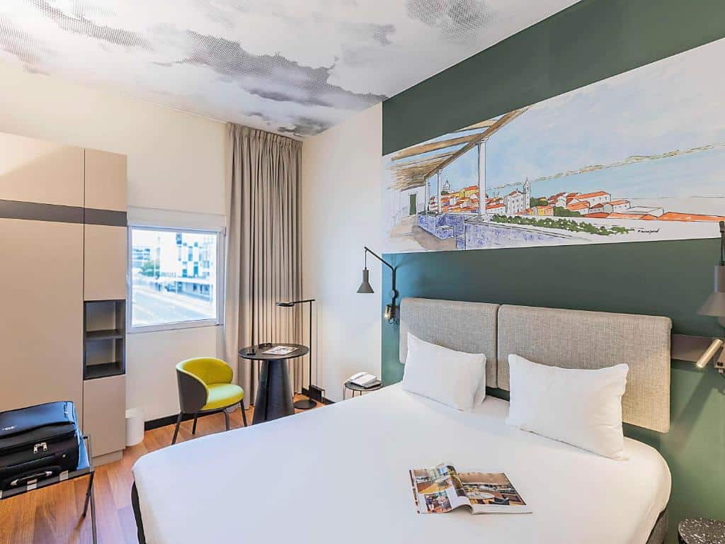 Quarto do Ibis Lisboa Parque das Nações com uma janela com cortinas, um pequeno armário, uma cama de casal, uma pequena mesinha com uma poltrona e há luminárias nas laterais da cama.