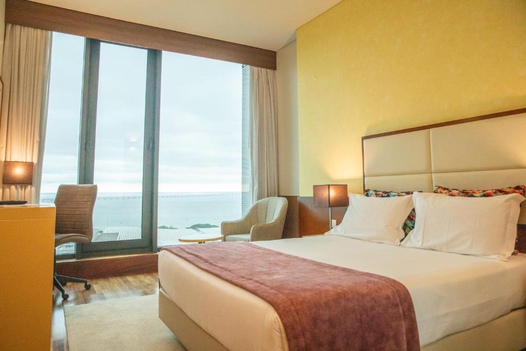 Um quarto moderno no VIP Executive Arts Hotel com uma cama de casal, escrivaninha, cadeira e uma grande janela com vista para o mar sob um céu nublado.