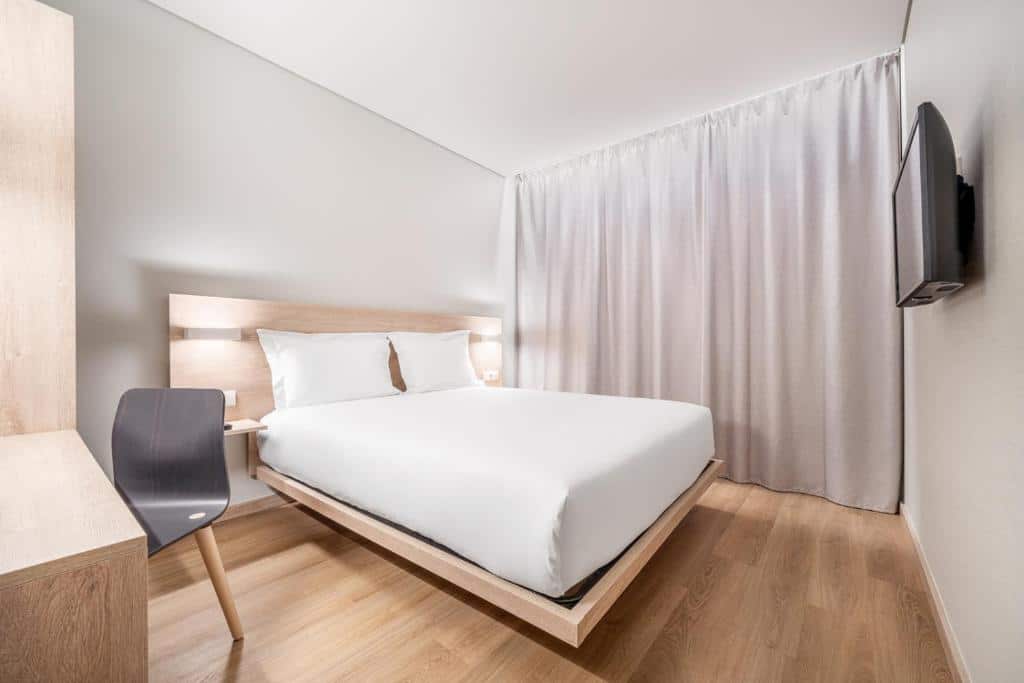 Quarto do Moov Hotel Lisboa Oriente com uma cama de casal, uma televisão de frente para a cama, uma mesa de escritório com uma cadeira e uma janela com cortinas.