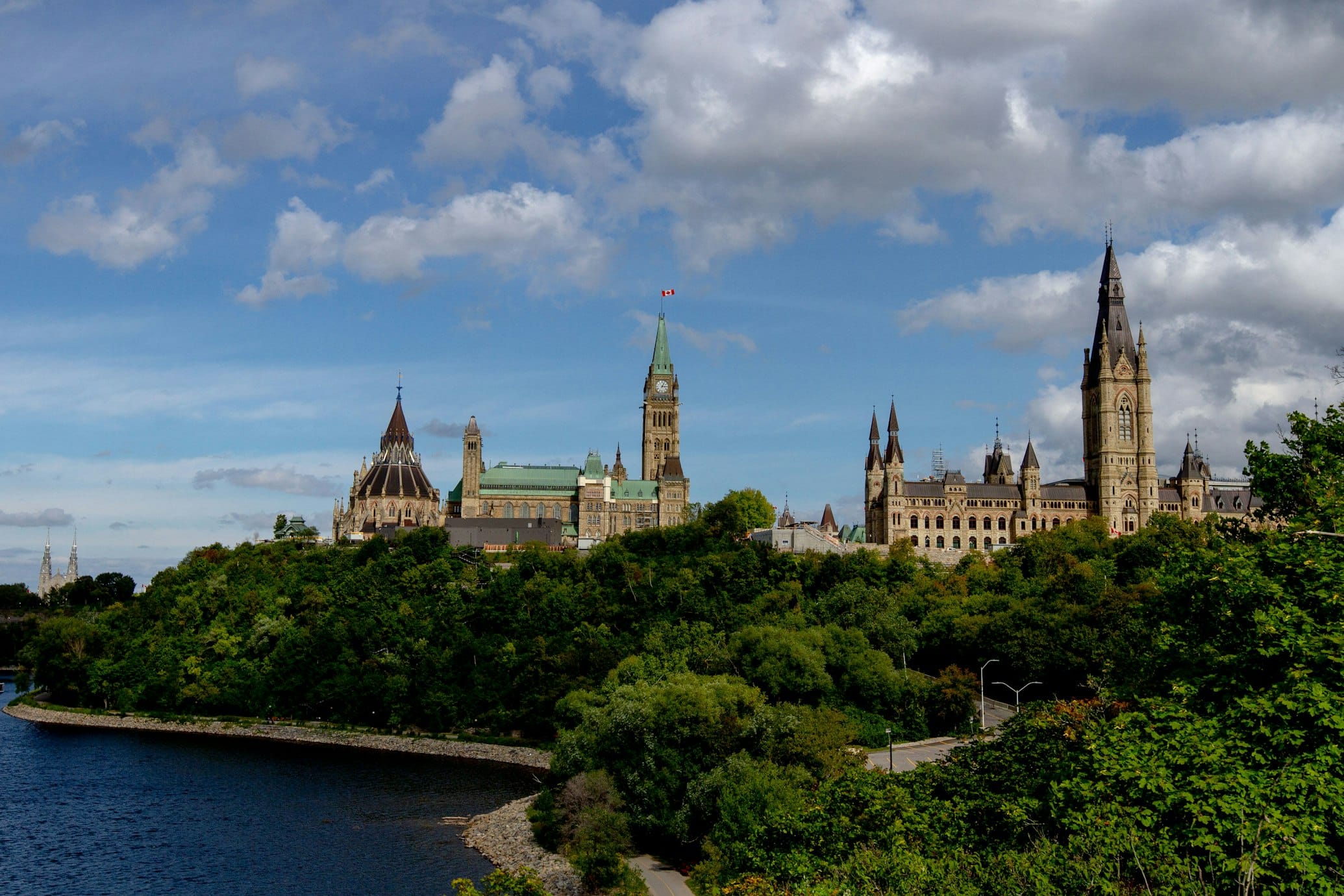 Vista da Colina do Parlamento do Canadá, com edifícios históricos cercados por árvores, ao lado de um rio sob um céu parcialmente nublado.
