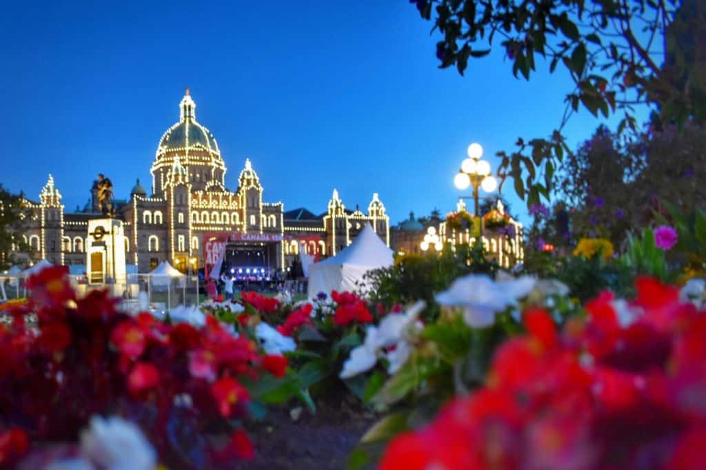 Edifício iluminado com uma cúpula à noite, com flores vermelhas e brancas em primeiro plano e árvores à direita. A imagem ilustra o post sobre intercâmbio no Canadá.