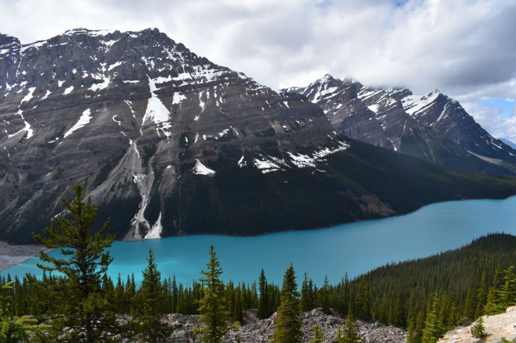 Um lago azul-turquesa cercado por árvores perenes com montanhas cobertas de neve e um céu nublado ao fundo. A imagem ilustra o post sobre intercâmbio no Canadá.