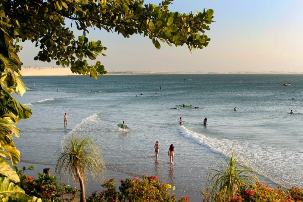 Foto da praia de Jericoacoara, com ondas baixas e pessoas se banhando e surfando. Perto da câmera há vegetação e vários arbustos com flores.