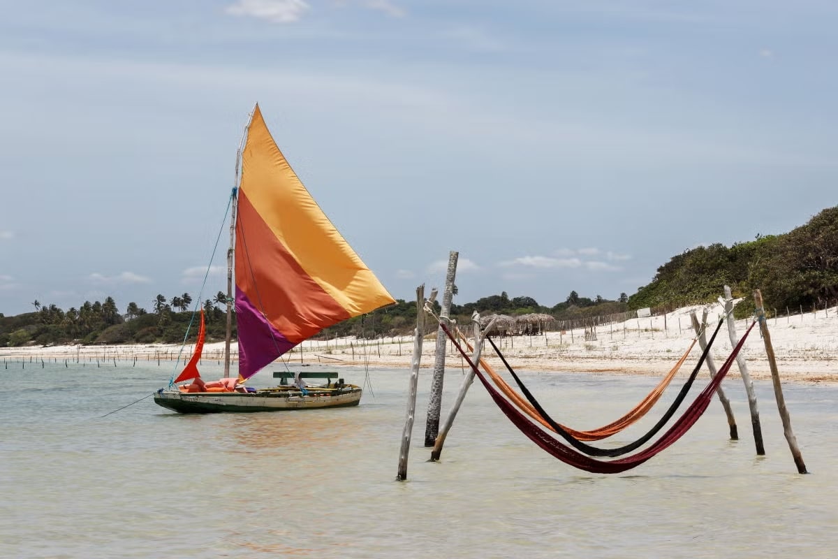 Foto mostrando a Lagoa do Paraíso. Vemos três e um barco com velas dentro da parte rasa da água. No fundo há a areia branca e a vegetação.