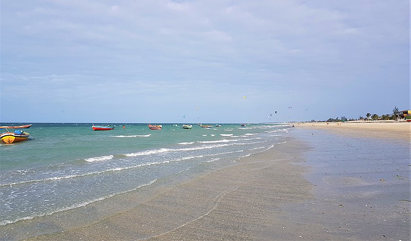 Foto na Praia do Preá, em Jericoacoara. Na direita está a areia e na esquerda, o mar. Há algumas canoas sobre a água. O céu é azul e com poucas nuvens.