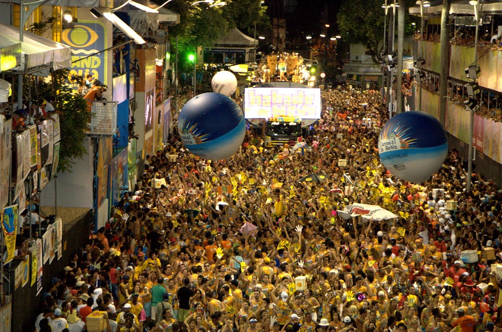 Uma grande multidão em Salvador comemora o Carnaval na rua à noite, com balões coloridos e luzes brilhantes, cercado por prédios altos.