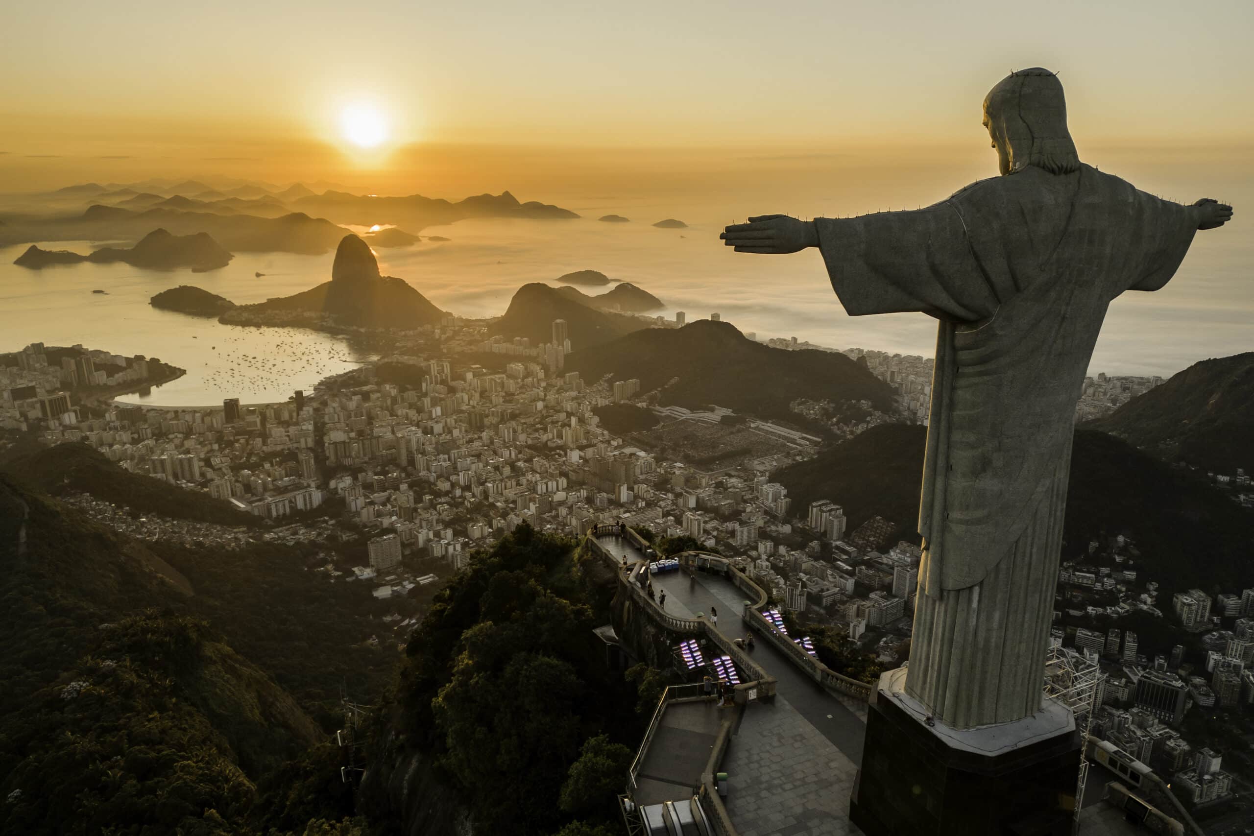 Vista aérea da estátua do Cristo Redentor com vista para o Rio de Janeiro ao pôr do sol, com o Pão de Açúcar ao fundo.