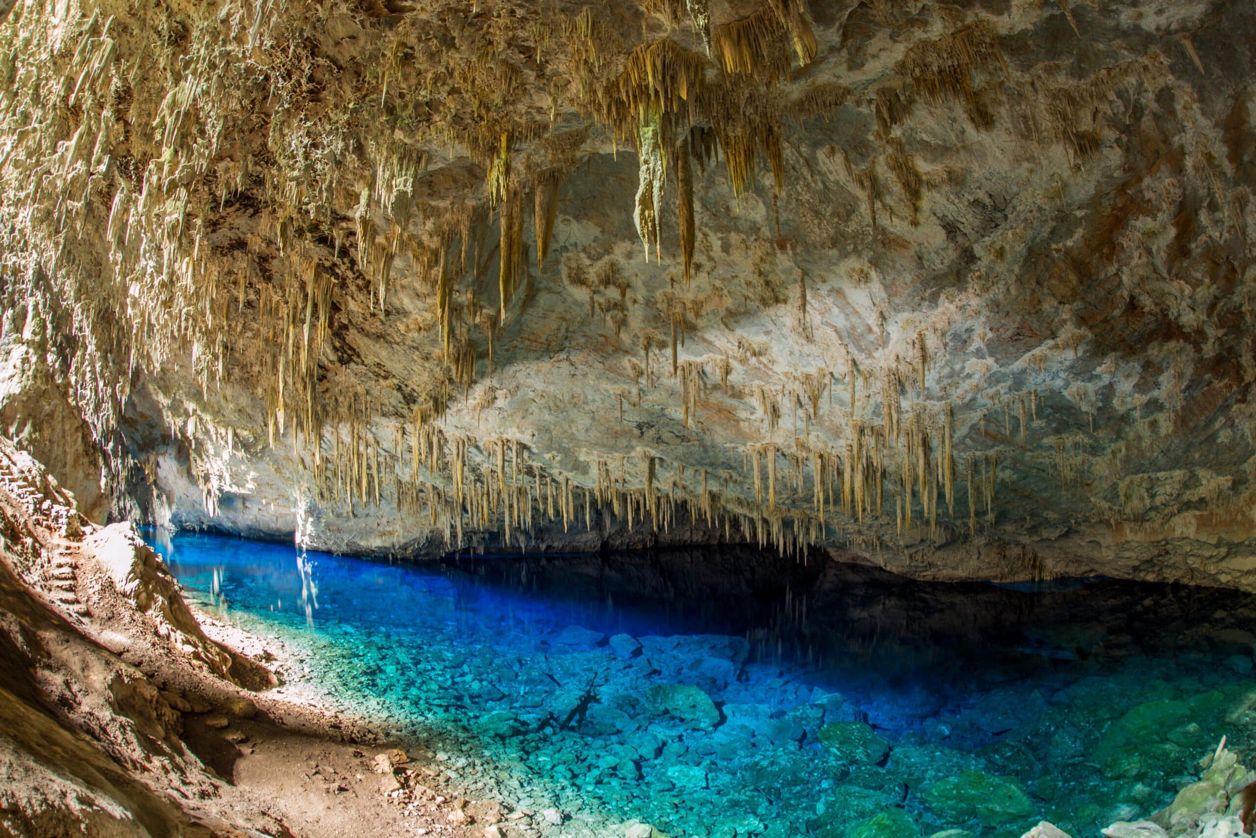 O interior da Gruta do Lago Azul com inúmeras estalactites penduradas no teto e uma piscina de água azul cristalina no chão rochoso.