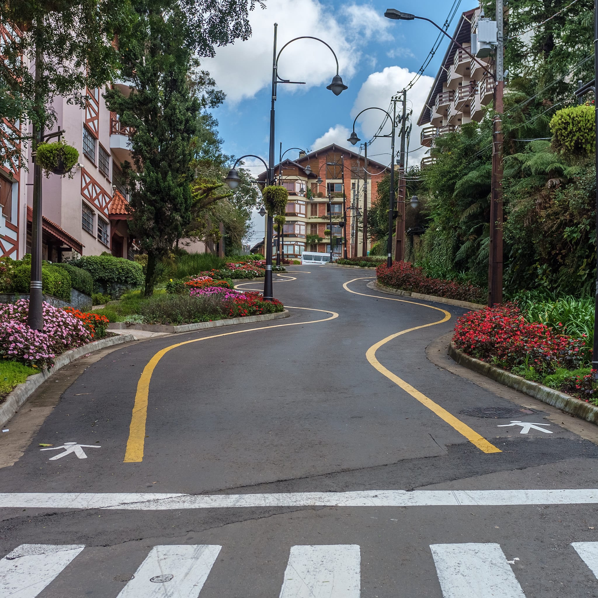 Estrada curva ladeada por flores e árvores coloridas, subindo em direção a edifícios sob um céu nublado em Gramado.