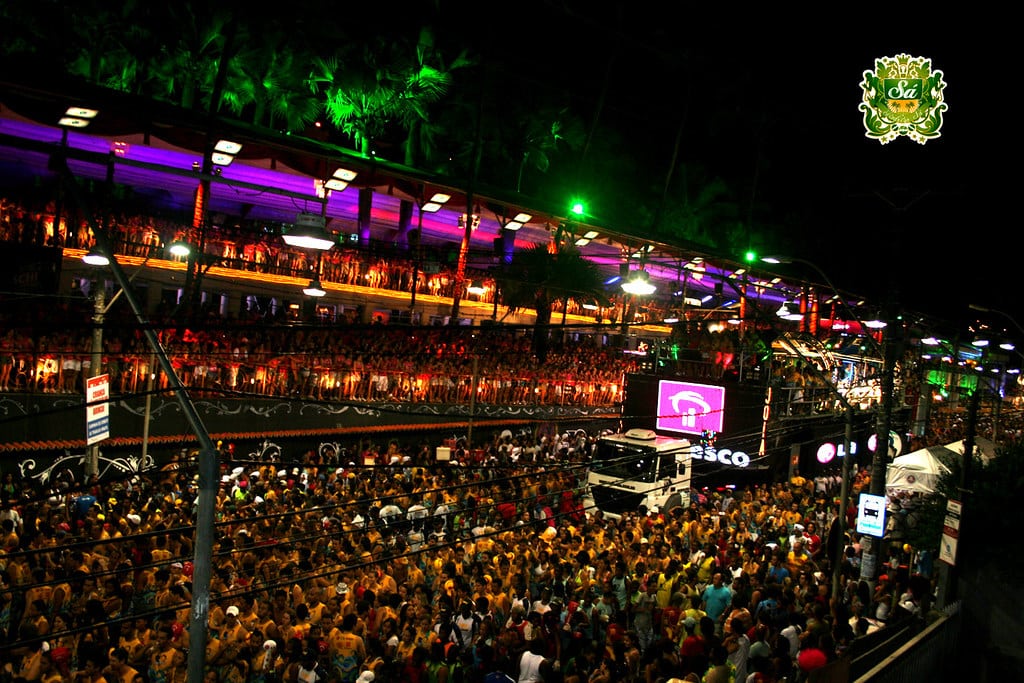 Uma grande multidão se reúne para o Carnaval de rua noturno em Salvador, com luzes coloridas e uma estrutura iluminada ao fundo.
