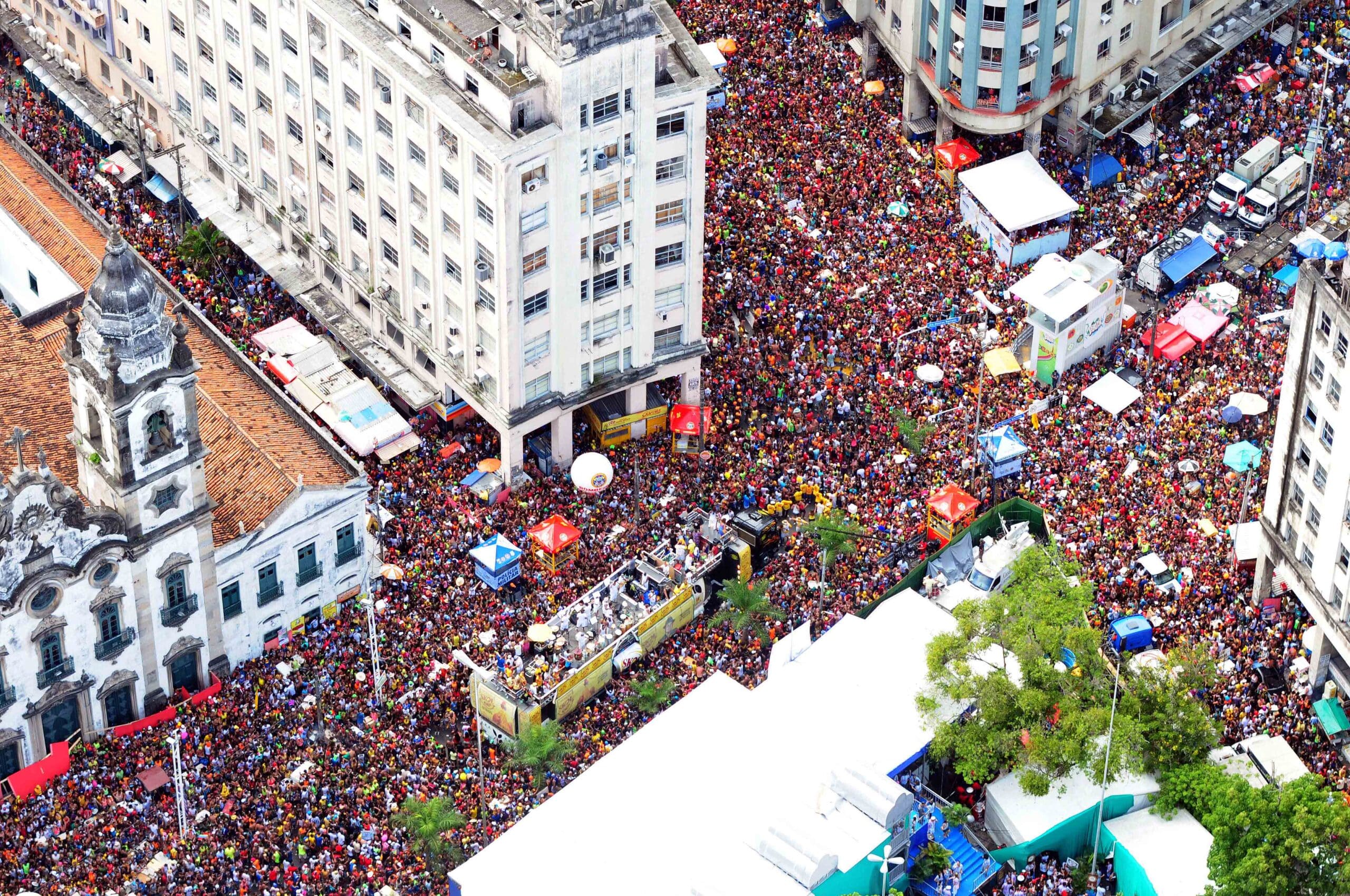 Vista aérea de uma grande multidão no Recife enchendo uma rua entre prédios altos e uma igreja em uma área urbana.