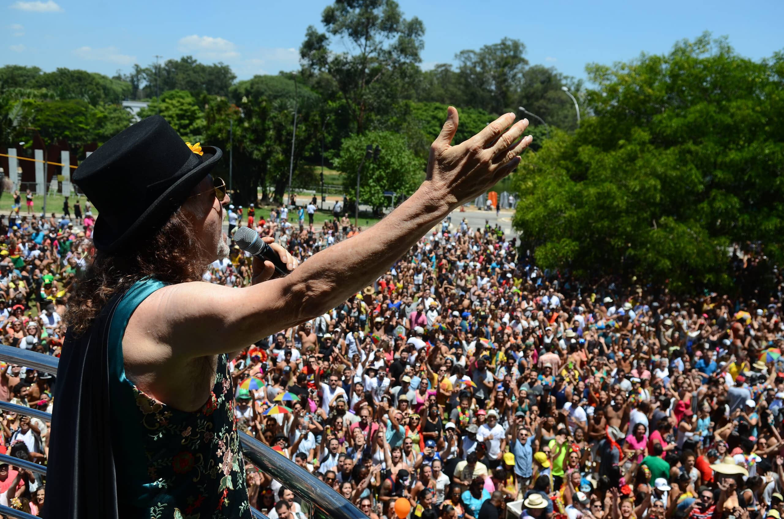 Pessoa com cabelo comprido e chapéu fala para uma grande multidão ao ar livre em um dia ensolarado em São Paulo.