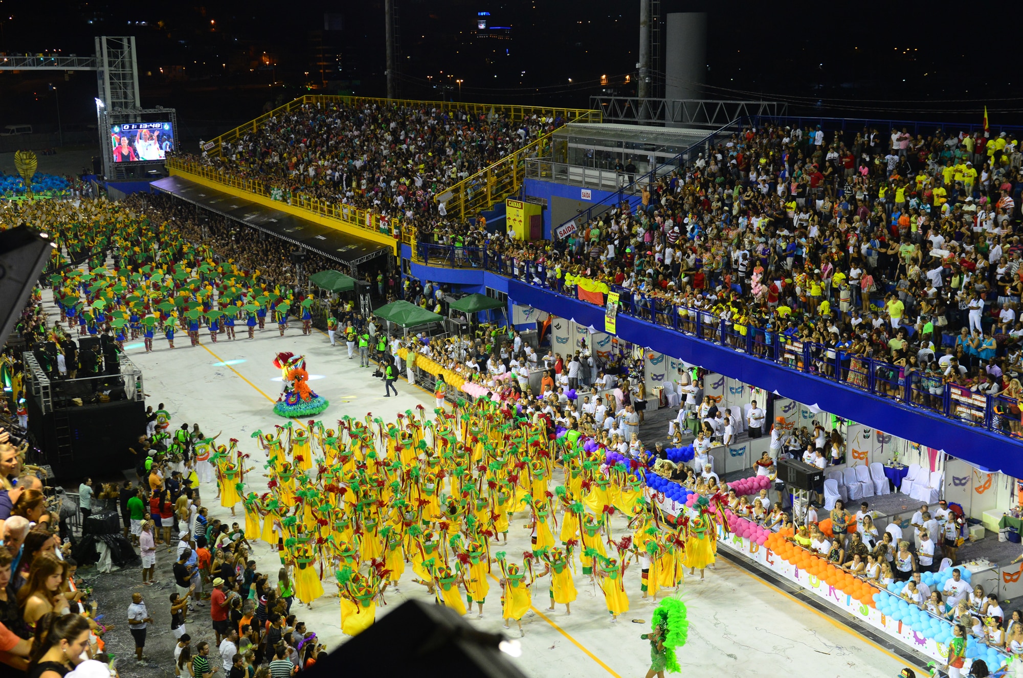 Uma grande multidão assiste a um desfile vibrante com dançarinos em trajes coloridos marchando por um amplo caminho em Florianópolis.