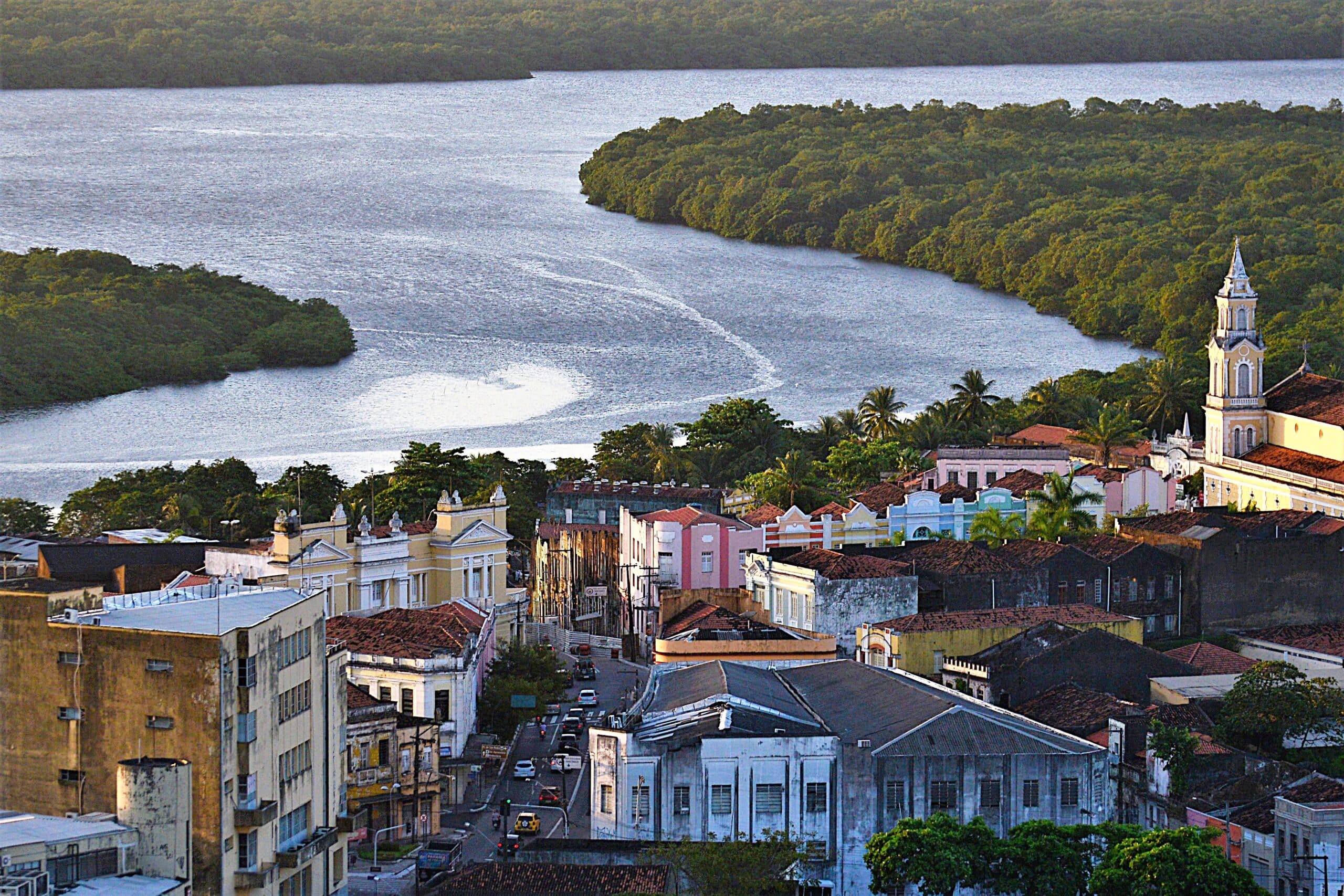 Vista aérea do Centro Histórico de João Pessoa com edifícios coloridos e uma igreja às margens de um rio, cercada por densa vegetação verde.