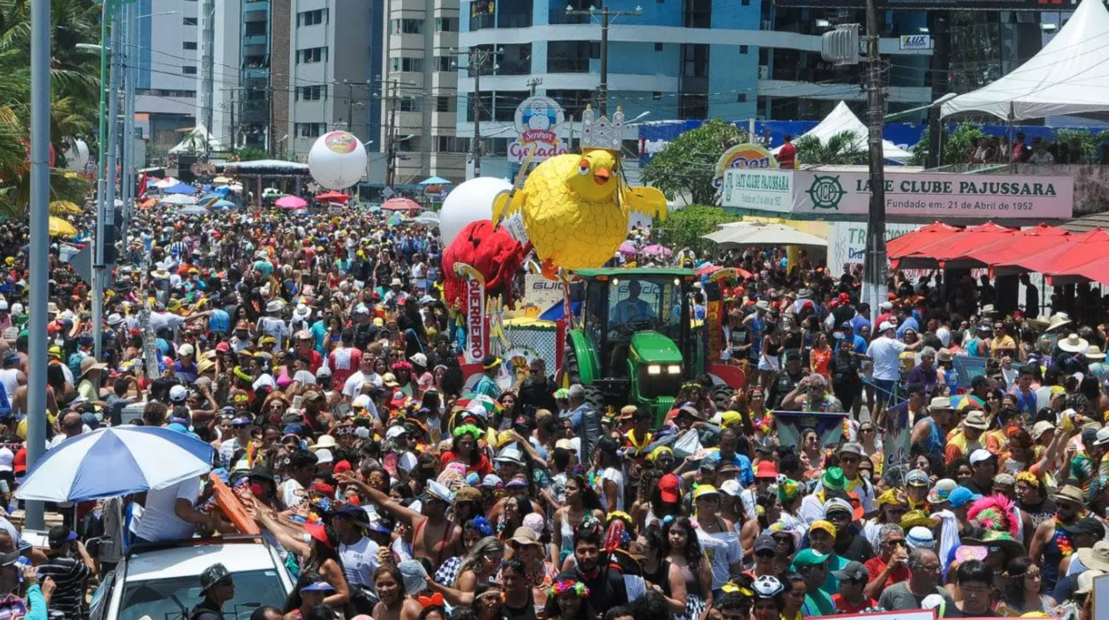 Um desfile grande e colorido com uma multidão de pessoas em trajes festivos em Maceió. Um carro alegórico com uma galinha amarela gigante é puxado por um trator.