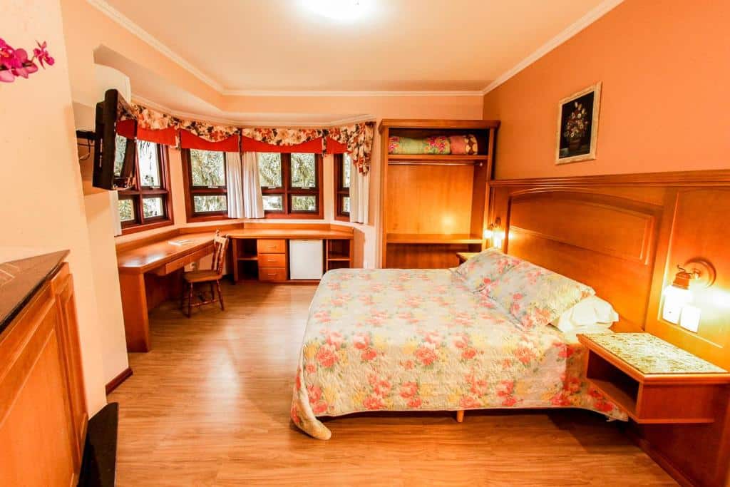 suíte da pousada das papoulas para representar onde ficar em Gramado. O quarto possui uma cama de casal à direita e uma mesa de madeira no canto esquerdo acompanhando toda a janela do espaço.