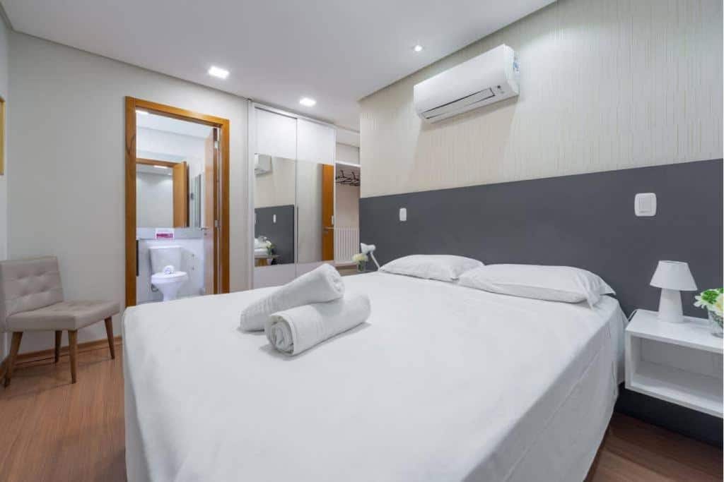 suíte do lindo apartamento de dois quartos no edifício paraíso mostrando uma cama de casal com duas toalhas brancas enroladas e colocadas em cima do colchão, uma porta aberta com acesso ao banheiro do quarto e um espelho ao fundo ao lado do armário embutido na parede.