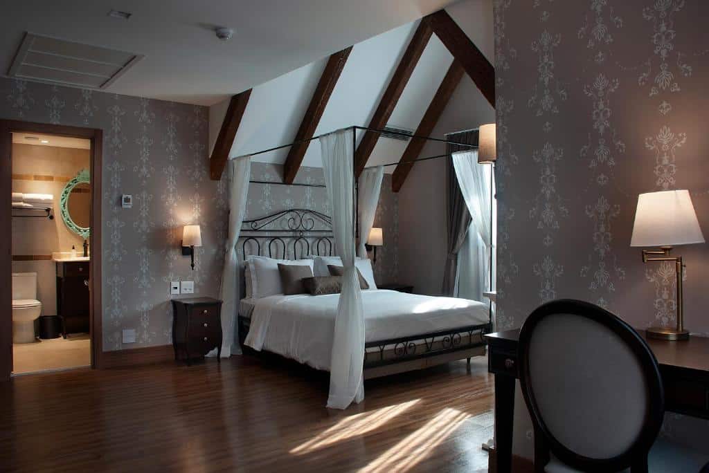 suíte do buona vitta gramado resort com uma cama de casal com dossel, vigas de madeira aparentes no teto, um porta à esquerda mostrando o acesso ao banheiro e uma cadeira acolchoada à direita, em frente a uma mesa de madeira com um abajur aceso em cima.